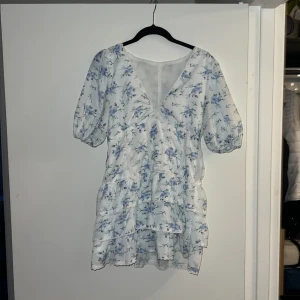 Blommig vit klänning med volanger - Har använt två gånger. Söt vit klänning med blått blommönster och volanger. Den har en v-ringad design och korta ärmar. Perfekt för sommarens alla tillfällen. Kan diskutera pris.