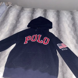 Mörkblå hoodie från Polo Ralph Lauren - Säljer en mörkblå hoodie från Polo Ralph Lauren med stor röd text 'POLO' på framsidan. Ärmen har en broderad amerikansk flagga och texten 'U.S.A.'. Perfekt för en avslappnad stil.