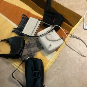 Samling av olika väskor - En samling av olika väskor inklusive en svart axelväska med spänne, en vit handväska med dragkedja, en rutig axelväska och en beige väska med krokodilmönster. Perfekt för olika tillfällen och stilar.