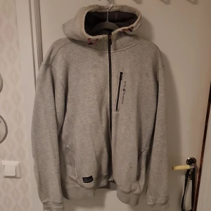 Grå huvtröja från Sail Racing - Säljer en grå huvtröja från Sail Racing med dragkedja och en bröstficka. Tröjan har en bekväm passform och är perfekt för kyligare dagar. Den har långa ärmar och en praktisk huva.
