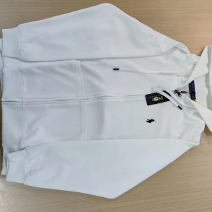 Vit hoodie från Polo Ralph Lauren - Snygg vit hoodie från Polo Ralph Lauren med dragkedja och broderad logga på bröstet. Perfekt för en avslappnad stil. Den har långa ärmar och en bekväm passform.