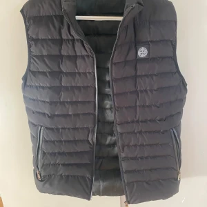 Svart dunväst med dragkedja M - Org pris 4000kr, Snygg svart dunväst från Stone Island med dragkedja och två praktiska fickor framtill. Västen har en quiltad design och en liten logotyp på bröstet. Perfekt för kyligare dagar.