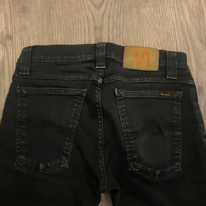 Svarta jeans från Nudie - Snygga svarta jeans från Nudie Jeans storlek 25/32// med en klassisk femficksdesign. De har en dragkedja och knapp framtill samt en läderpatch med logga på baksidan. Perfekta för en stilren look.de är avklippta längs ner så de passar folk som är 160-170. 