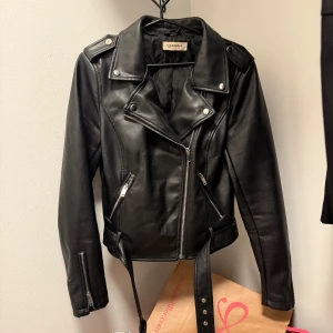 Svart skinnjacka från Glamorous - Snygg svart skinnjacka från Glamorous med dragkedja och bälte. Jackan har långa ärmar och stiliga detaljer som knappar på axlarna och dragkedjor vid ärmsluten. 