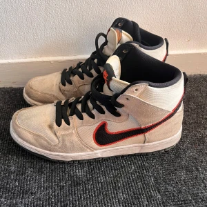 Nike Dunk High sneakers - Säljer ett par Nike Dunk High sneakers i beige mocka med svarta och röda detaljer. Skorna har en hög design med snörning och en rund tå. Perfekta för en stilren look. Skorna är INTE kopior alltså är de äkta!