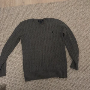 Grå kabelstickad tröja från Polo Ralph Lauren - Säljer en stilren grå kabelstickad tröja från Polo Ralph Lauren. Tröjan har långa ärmar och en diskret logga på bröstet. Perfekt för kyligare dagar. Storlek 18-20, passar bra M