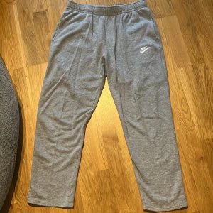 Grå mjukisbyxor från Nike - Gråa mjukisbyxor från Nike med elastisk midja och klassisk Nike-logga på vänster lår. Byxorna har raka ben, sidofickor och en bakficka med lock. Perfekta för chill eller träning.