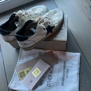 Golden Goose vita sneakers med svart stjärna - Golden Goose ballstar sneakers i vitt skinn med svart stjärna på sidan och svarta detaljer vid hälen. Skorna har snörning, rund tå och en vintage-inspirerad, krackelerad finish på hälen. Sulan är beige och har tydlig struktur undertill. Perfekta för dig som gillar en unik och trendig look.