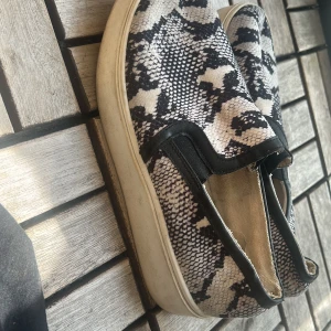 Slip-on sneakers med ormmönster - Svarta och vita slip-on sneakers med coolt ormmönster. Skorna har platt sula och elastiska partier vid öppningen för enkel påtagning. Ovandelen är i textil och detaljer i svart syntet. Perfekta för dig som vill sticka ut med din stil.