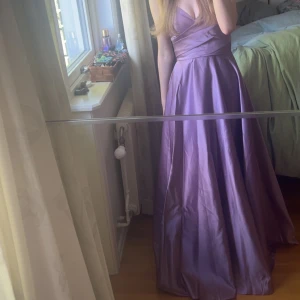 Lila balklänning med snörning i ryggen - Säljer en lila långklänning i glansigt material med v-ringad omlottliv och smala axelband. Klänningen har öppen rygg med snygg snörning och ett svepande, utsvängt kjolparti. Perfekt för bal eller student! Endast använd en gång på min bal i 9an så i superfint skick!