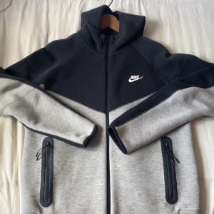 Nike svart och grå vindjacka - Nike vindjacka i svart och ljusgrått med hel dragkedja och hög krage. Jackan har två fickor med dragkedja och en sportig look. Perfekt för chill dagar eller när du vill ha en clean stil. Nike-logga på bröstet.