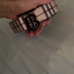 Burberry rutig bälte med B-spänne - Snyggt bälte från Burberry med klassiskt rutigt mönster i beige, svart och rött. Bältet har ett stort metallspänne med ett stilrent B i guld och svart. Perfekt accessoar för att lyfta din outfit med en lyxig touch.