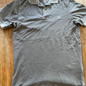Grå pikétröja från GANT - Klassisk grå pikétröja från GANT med broderad logga på bröstet. Tröjan har korta ärmar, krage och knäppning med två knappar framtill. Tillverkad i mjuk bomull som känns skönv mot huden. 