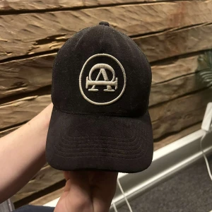 Svart keps från All Cap One - Svart keps från All Cap One med vit broderad logga framtill. Klassisk trucker-modell med mesh på sidorna och baksidan för extra ventilation. Justerbar passform och böjd skärm. Perfekt för en cool och avslappnad stil.
