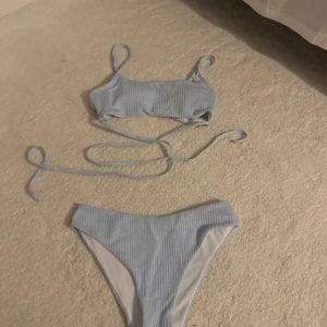 Ljusblå ribbad bikini med snörning - Superfin ljusblå bikini med ribbad struktur. Toppen har smala axelband och långa snören som kan knytas runt midjan för en snygg detalj. Bikinitrosan är högt skuren och matchar toppen perfekt. Perfekt för stranden eller poolen i sommar!
