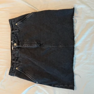 Svart jeanskjol från NA-KD - Snygg svart jeanskjol från NA-KD med rå kant nertill och klassiska fickor fram och bak. Kjolen har bälteshällor och knappstängning framtill. Perfekt för dig som gillar en enkel men edgy stil.