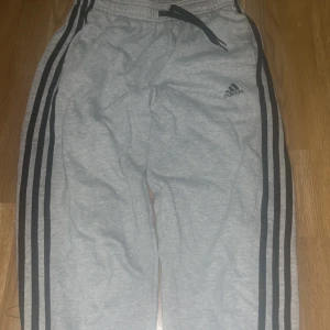 Grå Adidas mjukisbyxor med svarta ränder - Snygga grå mjukisbyxor från Adidas med klassiska svarta ränder längs sidorna och Adidas-logga på låret. Byxorna har resår i midjan och snörning för extra komfort. Perfekta för chill eller träning.