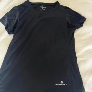 Svart träningst-shirt från Ronhill - Svart t-shirt från Ronhill i sportig modell, perfekt för träning. Gjord i ett lätt och snabbtorkande syntetmaterial med rund hals och korta ärmar. Diskret logga framtill nedtill. Sitter snyggt och är riktigt bekväm när du rör på dig. Endast testad. 