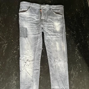 Dsquared2 jeans - Säljer ett par grå jeans från Dsquared2 med coola slitningar och patchdetaljer. Byxorna har knappar i gylfen, klassisk femficksmodell och en stor Dsquared2-patch bak. Snyggt tvättade och med en streetig vibe. Storlek 50, M och passar S och L , skriv för mer info 
