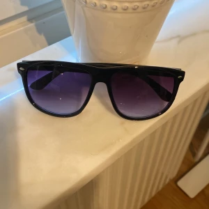 Rayband boyfriend likande solglasögon  - Snygga svarta solglasögon med klassisk wayfarer-inspirerad form och lila tonade glas. Bågarna är i blank plast och har små silvriga detaljer vid hörnen. Perfekta för att ge din look en cool vibe under soliga dagar. Aldrig använda🙌🤝