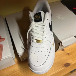 Nike Air Force 1 vit/svart/guld - Nike Air Force 1 sneakers i vitt skinn med svart swoosh och gulddetaljer. Klassisk låg modell med perforerad tå, snörning och vit sula. Guldmärke på snörningen och liten guldfärgad swoosh på sidan ger extra stilpoäng.