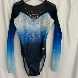 Blå glittrig gymnastikdräkt med stenar - Snygg långärmad gymnastikdräkt i blått och svart med glittriga stenar och coolt mönster. Materialet är stretchigt och består av 80% polyester och 20% elastan. Mycket lite använd.
