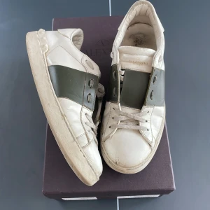 Valentino Garavani Open sneakers, vit/oliv - Snygga Valentino Garavani Open sneakers i vitt skinn med bred olivgrön panel över mitten. Klassisk låg modell med snörning och rund tå. Sulan har diskreta nitar längs kanten för en cool touch. Perfekta för dig som gillar exklusiva och stilrena sneakers. Sulan har nyligen blivit limmad fast det är inget man varken tänker på eller märker på skon.