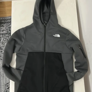 Grå och svart vindjacka från The North Face - Snygg grå och svart vindjacka från The North Face med huva och hel dragkedja. Jackan har en diskret logga på bröstet och ryggen, samt en praktisk ficka med dragkedja på armen. Passar max 180
