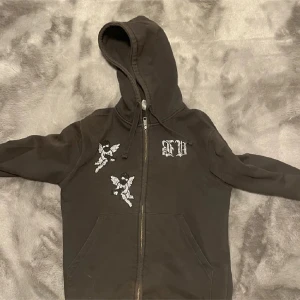 Svart hoodie med änglar och dragkedja - Svart hoodie med huva och dragkedja framtill. På bröstet finns två vita änglamotiv och ett tryckt bokstavstryck i vitt. Tröjan har känguruficka och snörning i huvan. Perfekt för en chill och stilren look.