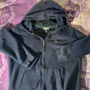 Mörkblå hoodie från Lacoste - Mörkblå hoodie från Lacoste med dragkedja och klassisk krokodillogga på bröstet. Tröjan har huva, långa ärmar och ribbade muddar. Perfekt för dig som gillar en clean och sportigh stil. Fickor framtill för extra chill vibe.