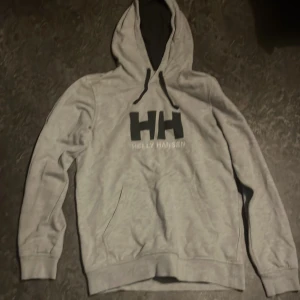 Grå hoodie från Helly Hansen M - Säljer en stilren grå hoodie från Helly Hansen i storlek M. Tröjan har svart HH-logga och broderad text på bröstet, huva med svarta snören och en klassisk känguruficka framtill. Perfekt för chill dagar eller när du vill ha en enkel streetlook.