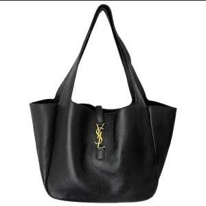 Svart shoppingväska från Saint Laurent - Stilren svart shoppingväska från Saint Laurent i mjukt skinn med guldigt YSL-emblem framtill. Väskan har rymlig design och långa handtag som gör den enkel att bära på axeln. Perfekt för dig som vill ha en lyxig och klassisk look.