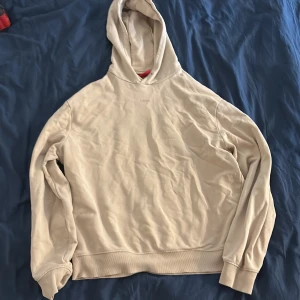 Beige hoodie från Hugo boss med huva - Säljer en stilren beige hoodie från Hugo med diskret logga framtill. Tröjan har huva och långa ärmar, samt ribbade muddar vid ärmslut och nederkant. Perfekt för en avslappnad och trendig look.