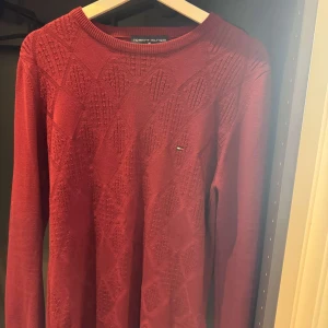 Röd stickad tröja Tommy Hilfiger - Snygg röd stickad tröja från Tommy Hilfiger med diskret logga på bröstet. Tröjan har ett subtilt mönster i tyget och rund halsringning. Perfekt för dig som gillar klassisk stil med en twist. Långärmad och mjuk i materialet.