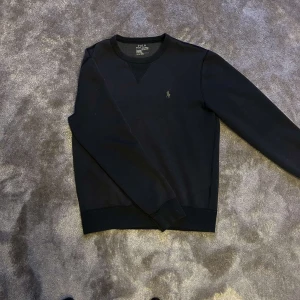 Svart sweatshirt från Polo Ralph Lauren - Svart sweatshirt från Polo Ralph Lauren med klassisk rund hals och broderad logga på bröstet. Tröjan har långa ärmar och ribbade muddar vid ärmslut och nederkant. Perfekt för en clean och stilren look.