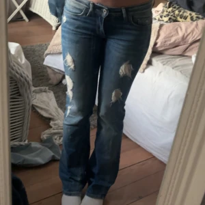 Lågmidjade jeans - Säljer ett par blå jeans med slitningar och hål på låren och knäna. Jeansen har bootcut passform, klassisk femficksdesign och lågmidja. Perfekta för en avslappnad och trendig look.