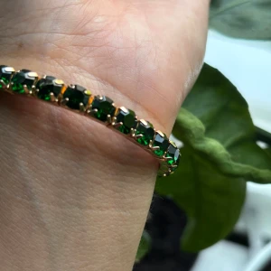Guldarmband med gröna stenar - Snyggt armband i guldton med glittrande mörkgröna stenar runt hela bandet. Stenarna är infattade i en klassisk tennisarmbandsstil som ger en lyxig och trendig känsla. Perfekt för dig som vill ha lite färg men ändå att det ska vara lite diskret och kunna matchas lätt med annat