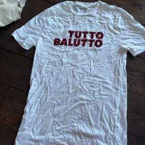 Vit T-shirt Tutto Balutto Nakata S - Vit t-shirt från Nakata med röd text 'TUTTO BALUTTO' tryckt på bröstet. Klassisk passform med rund hals och korta ärmar. Tillverkad i mjuk bomull, perfekt för dig som gillar sportiga och stilrena plagg med en twist.