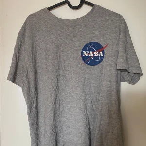 Grå NASA t-shirt  - Grå t-shirt från H&M med klassiskt NASA-tryck i blått, vitt och rött både fram och bak. T-shirten är kortärmad och har en avslappnad passform. Tillverkad i mjuk bomull, perfekt för dig som gillar rymd och coola prints. Frakt tillkommer
