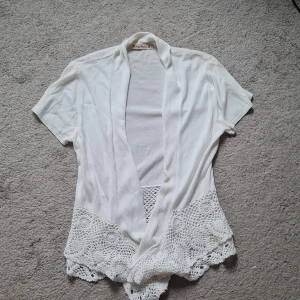Vit kofta med spets från Vintage Stories - Supersöt vit kofta från Vintage Stories med korta ärmar och öppen front. Nedre delen har snygga virkade spetsdetaljer som ger en boho-vibe. Perfekt att slänga över en topp för en chill och stilren look. Står storlek 40/42 men passar på mig som har xs. Då sitter den lite mer avslappnat och ned till höfterna.
