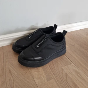 Svarta sneakers från Hartal med dragkedja - Svarta sneakers från Hartal med chunky sula och dragkedja framtill. Ovandel i syntetmaterial med detaljer i skinnimitation och rund tå. Snygg och stilren design med diskret logga på plösen. Perfekta för dig som gillar minimalistisk och sportig look. 