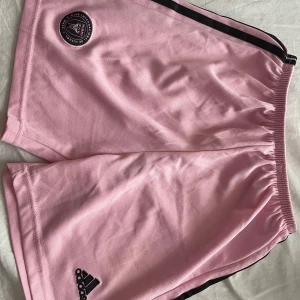 Rosa Inter Miami shorts från Adidas - Säljer ett par ljusrosa Inter Miami shorts från Adidas med svarta detaljer och klubbmärke på ena benet. De har elastisk midja och är gjorda i lätt och ventilerande polyester, perfekt för fotboll eller träning. Klassisk Adidas-design med svarta ränder längs sidorna. Tyvärr har numret fallit av. 
