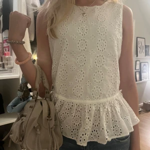 Vit broderad blus med volang - Supersöt vit blus med broderade blommor och volang nertill. Ärmlös modell i luftig bomull, perfekt för varma dagar. Ger en romantisk och fräsch känsla till outfiten.