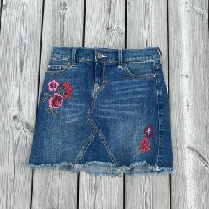 Blommig jeanskjol Abercrombie Kids - Supersöt jeanskjol från Abercrombie Kids med broderade blommor i rött och rosa framtill. Kjolen har fransig nederkant, klassiska fickor och snygga detaljer på bakfickorna. 
