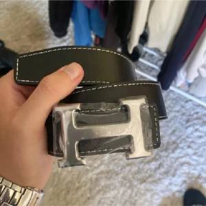 Svart skärp med H-spänne - Snyggt svart bälte i skinn med vit kontrastsöm och ett stort, silvrigt H-format spänne. Perfekt accessoar för att lyfta vilken outfit som helst med en clean och modern vibe.