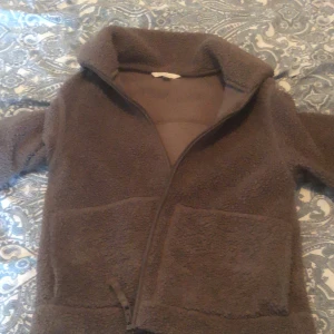 Brun teddyjacka från H&M - Mysig brun teddyjacka  med dragkedja framtill och stora fickor. Jackan har en hög krage och är tillverkad i ett fluffigt teddy-material som håller dig varm under kyliga dagar. Perfekt för dig som gillar en chill och avslappnad stil.