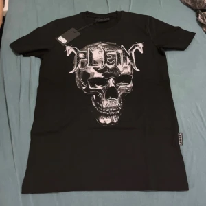 Svart t-shirt med dödskalle Philipp Plein - Svart t-shirt från Philipp Plein med stor silvrig dödskalle och logga framtill. T-shirten har korta ärmar, rund hals och coola detaljer med blanka nitar runt trycket. Materialet är mjuk bomull och passformen är normal.