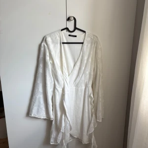 Vit omlottklänning med blommönster från Gina Tricot - Superfin vit kort omlottklänning från Gina Tricot  med subtilt blommönster i tyget. Klänningen har långa ärmar, v-ringning och knytband i midjan. Används en gång på skolavslutning. Nästan helt nytt skick. 