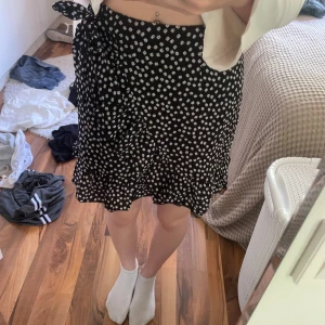 Svart blommig omlottkjol från SHEIN - Supersöt svart kortkjol från SHEIN med vita små blommor över hela tyget. Kjolen har omlottmodell med knyt i sidan och volangkant nertill som ger en extra feminin vibe. Perfekt för dig som gillar en lätt och flowy stil. Bara testad