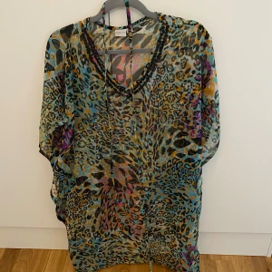 Färgglad leopardmönstrad kaftanblus - Säljer en luftig och transparent kaftanblus med leopardmönster i blått, gult, svart och lila. Blusen har v-ringning, knytband att knyta runt midjan och lös passform – perfekt för sommardagar eller strandhäng.